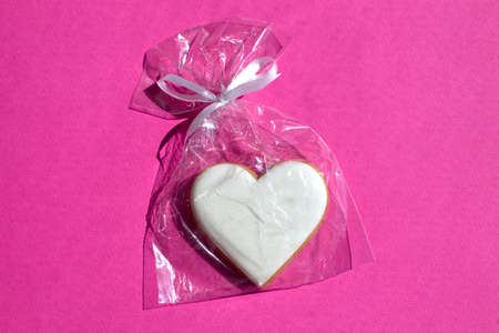 Gingerbread heart decorated with icing in gift plastic bag with ribbon on bright pink background...