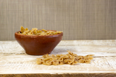 Whole grain dark pasta bows on brown plate on a rough wooden light background with selected focus