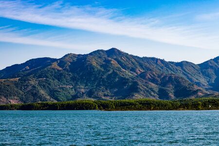 A lake with blue water around which grows green trees in the forest, and behind it high mountains. In the background is a blue sky with white cloudsの写真素材