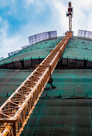 A construction crane near the building under construction, diagonally constructed of yellow parts and surrounded by a green protective grid. Against the background of blue sky and cloudsの写真素材