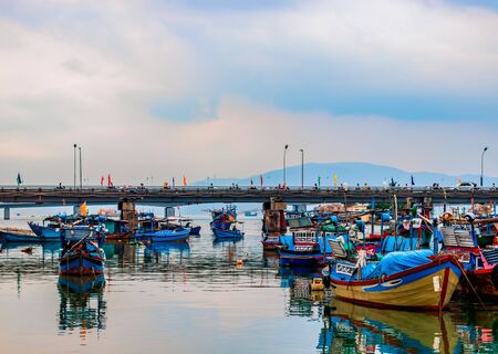 Fisherman boats on the coast of the riverwith reflection in the water. In the background there is a bridge where people ride mopeds and the sky with cloudsの写真素材