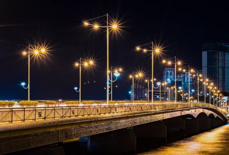 Concrete road bridge over the river with glowing orange lights on both sides. In the background are high-rise buildings of the city. At a slow shutter speedの写真素材