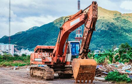 An orange excavator stands on a road with a bucket lowered near a landfill. In the background a high mountain and an antennaの写真素材