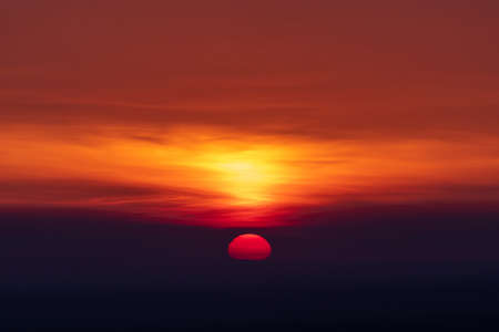 The silhouette of the sun over the horizon illuminates the clouds. Below the horizon is darknessの写真素材