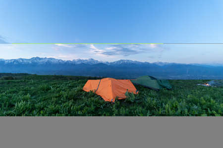 Two tents stand at the top of a green hill. In the background mountain peaks and cloudsの写真素材