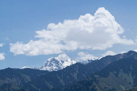A snowy mountain peak behind the hills and white clouds above it. Against the blue skyの写真素材