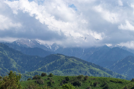 View of the green hills and clouds that cover the peaks of the snowy mountainsの写真素材