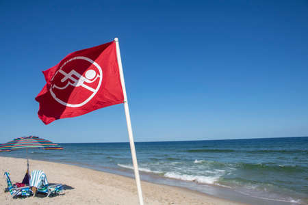 Red and white no swimming symbole icon on a beach flag spiked into the beach sand with ocean water and waves behind itの写真素材