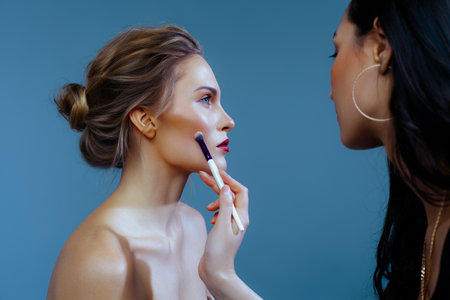 Makeup artist applies blush with brush to face of beautiful blonde on blue background. backstage work, workflowの写真素材