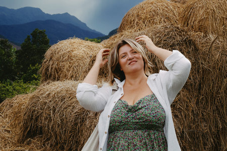 Adult caucasian female smiling by haystacks with mountainous backdrop.