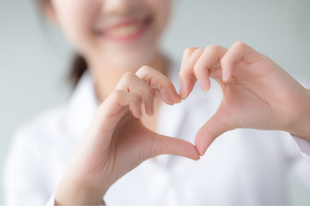 A smiling young woman dressed in a white shirt holds up her hands, forming a heart shape with her fingers. She is indoors against a light, neutral background.の素材