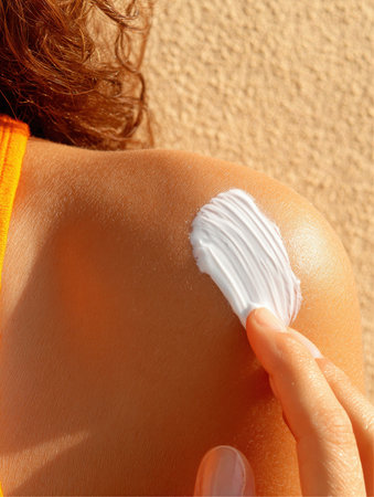 A woman is applying white sunscreen on her shoulder. She is wearing a yellow swimsuit, and the sun is shining brightly during the day, likely at a beach location.の素材