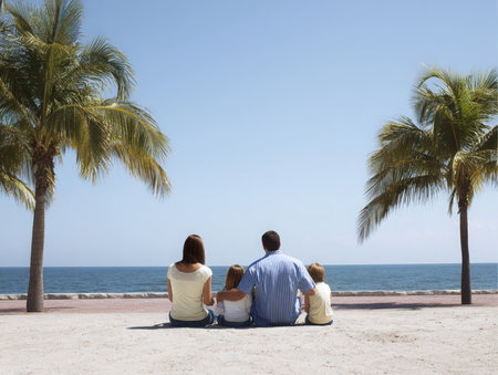 A family of four, a mother, father, daughter, and son are sitting on a sandy beach, looking out over the ocean in Puerto Vallarta, Mexico. Two palm trees stand on either side of them.の素材