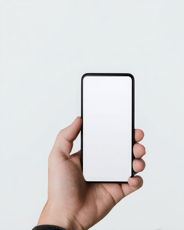 A hand holds a mobile telephone vertically. The cell phone has a black frame, and a blank, white screen. The background is solid white.の素材