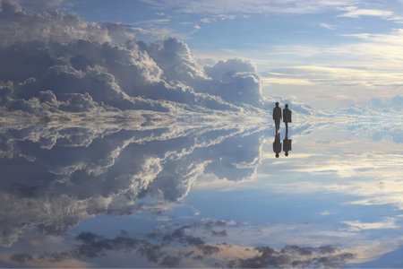 Two people walk on a reflective plain surface under a bright sky with many clouds. The ground appears to mirror the sky, creating a surreal effect in Uyuni, Bolivia.の素材