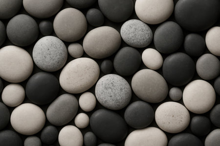 A detailed perspective captures a collection of smooth, rounded river pebbles. The stones, varying in size from small to medium, are tightly interlocked, creating an organic and consistent pattern across the entire frame. Their natural color palette ranges from deep black to various shades of grey and soft cream, offering a subtle contrast that enhances the textural depth. Each pebble exhibits a polished, matte finish, suggesting years of natural erosion. This arrangement evokes a sense of calm and natural simplicity, perfect for backgrounds, relaxation themes, or design elements requiring an earthy, grounding aesthetic.の素材