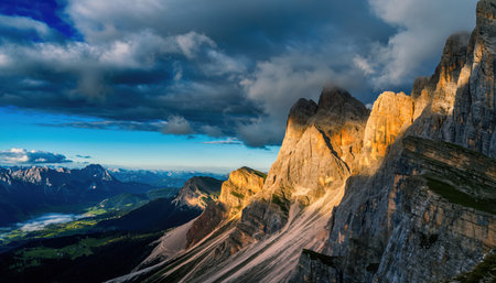 An expansive view of rugged mountain peaks bathed in warm golden light, likely during sunrise or sunset. Dark, dramatic clouds fill the sky, contrasting with patches of bright blue. In the distant valley, green fields and smaller mountains are visible, creating a majestic alpine panorama.の素材