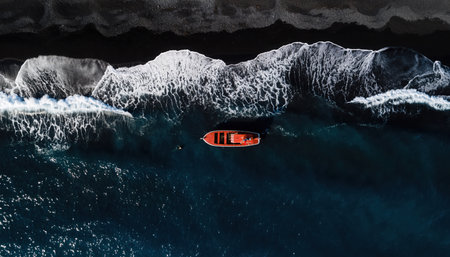 An aerial perspective captures a small, reddish-orange boat, possibly a kayak or paddleboard, floating in the vast, deep blue-green ocean water. White foamy waves are breaking near a dark, rocky shoreline at the top of the frame. The scene conveys a sense of solitude and the expansive power of nature.の素材
