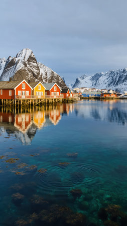 Traditional red and yellow Norwegian rorbuer cabins on stilts reflect in clear blue fjord water. Majestic snow-capped mountains rise behind them under a bright sky, showcasing a serene coastal landscape.の素材