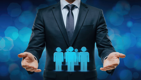 The photograph features a professional individual, depicted from the chest down, wearing a dark business suit and a white shirt with a tie. Their hands are open and extended forward, palms upward, creating a supportive gesture. Above their hands, three luminous, cyan-colored silhouette figures, representing individuals or a group, appear to float. These figures are stylized, showing distinct outlines for a male and two female forms. The background is a dynamic, blurred expanse of blue, punctuated by subtle, interconnected hexagonal light patterns, evoking a sense of digital technology, data, or interconnectedness. This visual metaphor conveys concepts such as human resource management, team support, community building, customer care, or the protection of a workforce within a technological or corporate environment. The composition emphasizes care, connection, and responsibility in a modern business context.の素材