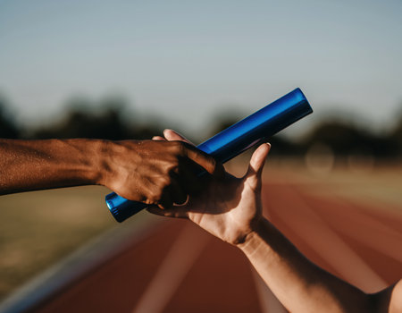 Close-up of two athletes hands exchanging a blue relay baton on a running track during a race. The focus is on the crucial moment of the handover, symbolizing teamwork, collaboration, and shared effort in athletics.の素材
