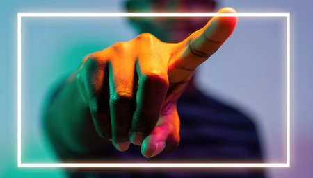 A close-up view of a persons outstretched hand with an index finger pointing directly towards the viewer. The hand is illuminated by vivid, colorful light, casting an orange glow on the skin. A blurred figure is visible in the background. A prominent white, luminous rectangular frame is superimposed, encompassing the pointing hand, suggesting a digital interface, selection, or interactive element. The scene evokes themes of technology, virtual reality, augmented reality, selection, and future interaction.の素材