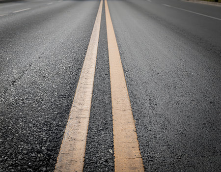 Close-up perspective of a worn asphalt road featuring two distinct, parallel solid yellow lines extending into the distance. The textured surface of the pavement is clearly visible under natural light.の素材