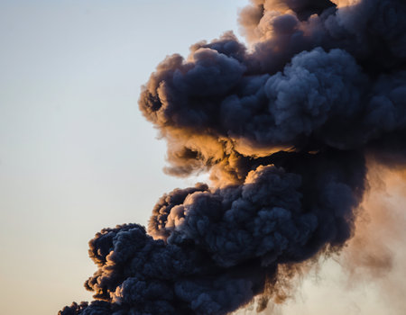 A thick, dark plume of smoke billows upwards against a light, clear sky. The smoke is dense and black, suggesting a large fire, industrial emission, or an environmental event. The sky has a subtle warm hue, possibly indicating dawn or dusk. The scene conveys a sense of pollution, disaster, or industrial activity impacting the atmosphere.の素材