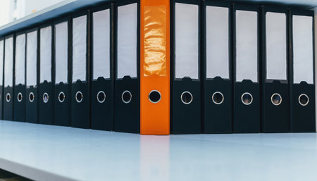 A single vibrant orange document folder stands out among a neat row of identical black binders on a clean, light shelf. This visual represents organization, important records, and distinctiveness within a corporate or administrative setting.の素材