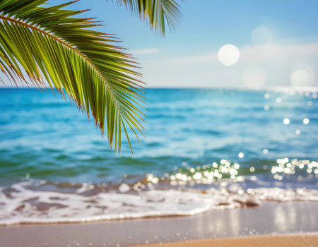 Gentle blue ocean waves meet a sandy tropical beach. A vibrant palm frond frames the view, with sparkling sunlight reflecting on the water under a clear sky.の素材