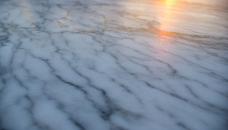 A detailed close-up reveals a textured frozen surface, showcasing intricate rippled patterns of ice or compacted snow. A soft, warm light from a distant sun, likely a sunrise or sunset, reflects upon the surface, creating a gentle golden glow and a subtle lens flare effect in the upper right. The scene depicts a serene and cold natural environment.の素材