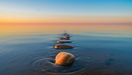 A breathtaking view of a tranquil seascape at either sunset or sunrise. The calm, reflective surface of the blue ocean water is dotted with a gentle line of smooth, rounded pebbles or small rocks, forming a natural path extending towards the distant horizon. The sky displays a beautiful gradient of colors, transitioning from warm oranges and soft pinks near the suns position to cool, clear blues higher above. The overall atmosphere is one of profound peace and serenity, making it ideal for concepts related to mindfulness, escape, or natural beauty. The subtle ripples around the submerged elements add texture to the otherwise smooth water.の素材