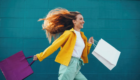 A cheerful young woman with vibrant, flowing curly hair runs enthusiastically against a solid teal background. She is smiling broadly, expressing pure joy and excitement. Dressed in a bright yellow jacket, a white top, and light green trousers, she carries two paper shopping bags, one purple and one white, suggesting a successful retail excursion. Her dynamic pose and beaming face convey happiness and energy, indicative of a pleasant consumer experience or simply enjoying lifes moments. The sharp contrast with the simple background highlights her movement and lively spirit.の素材