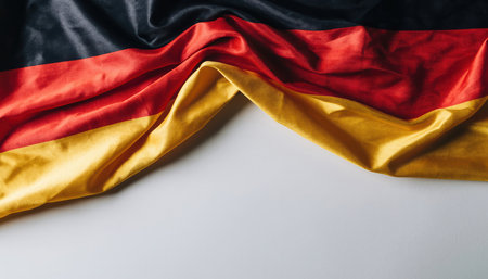 A close-up of a vibrant German flag, its black, red, and gold fabric subtly crumpled, draped across a pristine light grey surface. The lower portion offers clear space for text.の素材