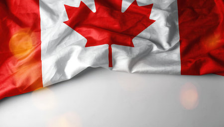 A vibrant Canadian flag is displayed, featuring the iconic red and white colors and the central maple leaf symbol. The fabric appears slightly worn and wrinkled, adding texture. It is draped across the top portion of a bright, light-colored background, which includes subtle bokeh light effects. This visual conveys themes of patriotism, national pride, and Canadian identity, suitable for various national events or cultural projects.の素材