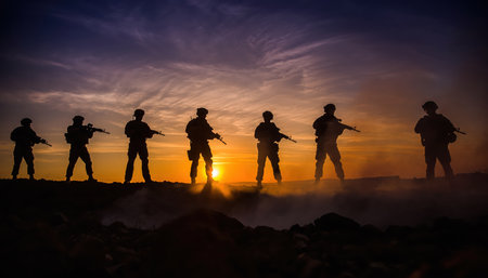 A group of seven silhouetted military personnel stand in a line on a low ridge or horizon against a vibrant sunset. The sun, a bright orange, is setting behind them, casting long shadows and creating a dramatic sky with hues of orange, purple, and dark blue. Each figure is carrying a rifle or firearm, suggesting an armed unit. Wisps of fog or smoke rise from the ground in front of them, adding to the intense atmosphere. The scene evokes concepts of defense, operation, security, and armed conflict.の素材
