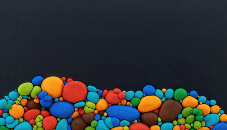 A close-up view of numerous small, multicolored, rounded elements forming a dense pile at the bottom of the frame. The elements display a variety of bright colors including red, blue, green, yellow, and orange, against a stark, dark background that occupies the upper portion, offering ample space for text or design. The texture appears smooth and uniform, creating an engaging visual pattern.の素材