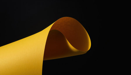 A close-up view of a vibrant golden yellow material, possibly paper or fabric, curled into a gentle arc. The material has a subtle textured surface and is brightly lit, contrasting sharply against a deep, dark black background. The composition is minimalist and abstract, highlighting the curve and the rich color. The focus is on the material's form and the interplay of light and shadow on its surface. No other objects or subjects are present, emphasizing simplicity and space.の素材