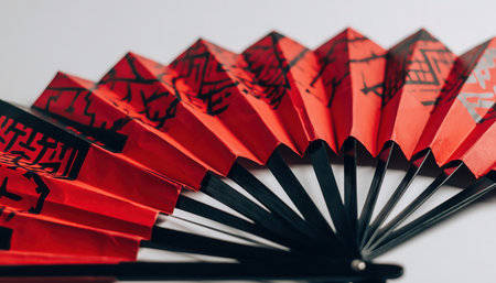 A partially opened traditional folding hand fan prominently displays its vibrant red segments, embellished with intricate black patterns. Its dark structure contrasts against a minimalist, light background, highlighting the cultural design.の素材