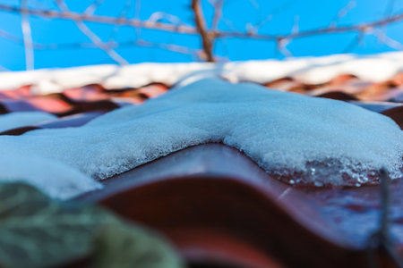 Soft white snow blankets a red tile roof, glistening under the bright blue sky. This tranquil winter landscape captures the essence of a serene season filled with beauty and calmness.の写真素材