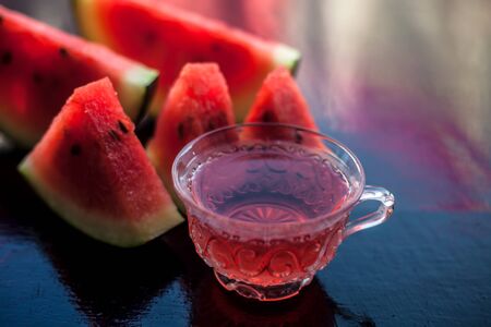 Mouth-watering watermelon tea in a transparent glass cup on wooden surface with watermelon pieces in triangle shape.の写真素材