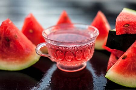 Fruit ice tea of watermelon seeds in a transparent glass cup on wooden surface along with triangular pieces of watermelon.の写真素材