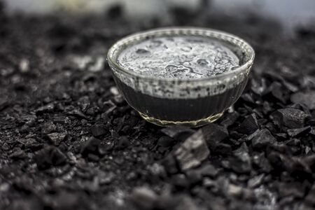 Close up of activated charcoal in a glass bowl on the wooden surface along with some raw powder of charcoal or coal spread on the surface.の写真素材