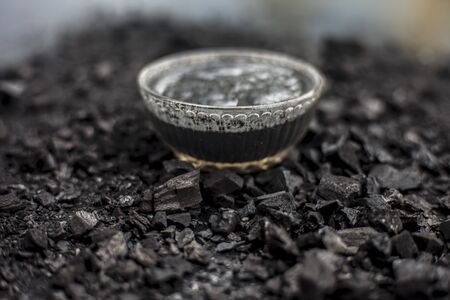 Close up of activated charcoal in a glass bowl on the wooden surface along with some raw powder of charcoal or coal spread on the surface.の写真素材