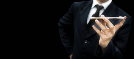 Young businessman or entrepreneur wearing a dark blue coat or jacket with a black necktie and blue shirt isolated on black with a cellphone in his hands.Horizontal shot.の写真素材