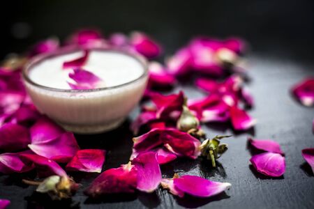 Best and affordable home face mask i.e. milk or milk cream with rose petals in a glass bowl on the black wooden surface along with some sprinkled rose petals.Horizontal shot.の写真素材