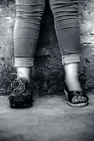 The elegant foot of young teen girl wearing peep-toe sandals in one foot and hiking boots in another posing against a brick wall wearing blue denim jeans. Vertical shot.の写真素材