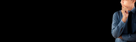 A young male worker dressed in a blue shirt and black necktie with his hand at his chin thinking something against a black background.の写真素材