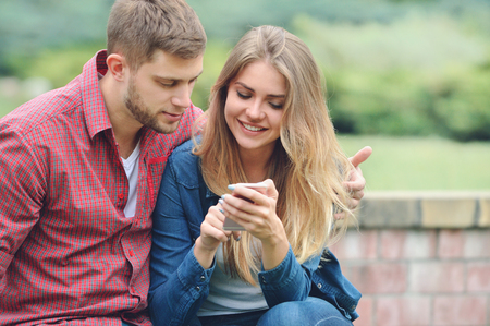Young couple looking at a smartphone together on a bench in the park. Shot of a happy couple browsing together on the internetの写真素材
