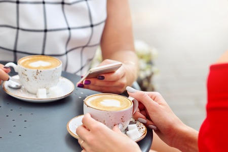 Close up view of two coffee cups and hands of young women with smartphone at outdoor cafe. Lifestyle, friendship and technology conceptの写真素材